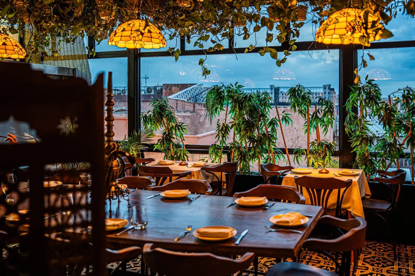 Chic restaurant interior featuring tables set for dining amid lush greenery, with cityscape visible through large window