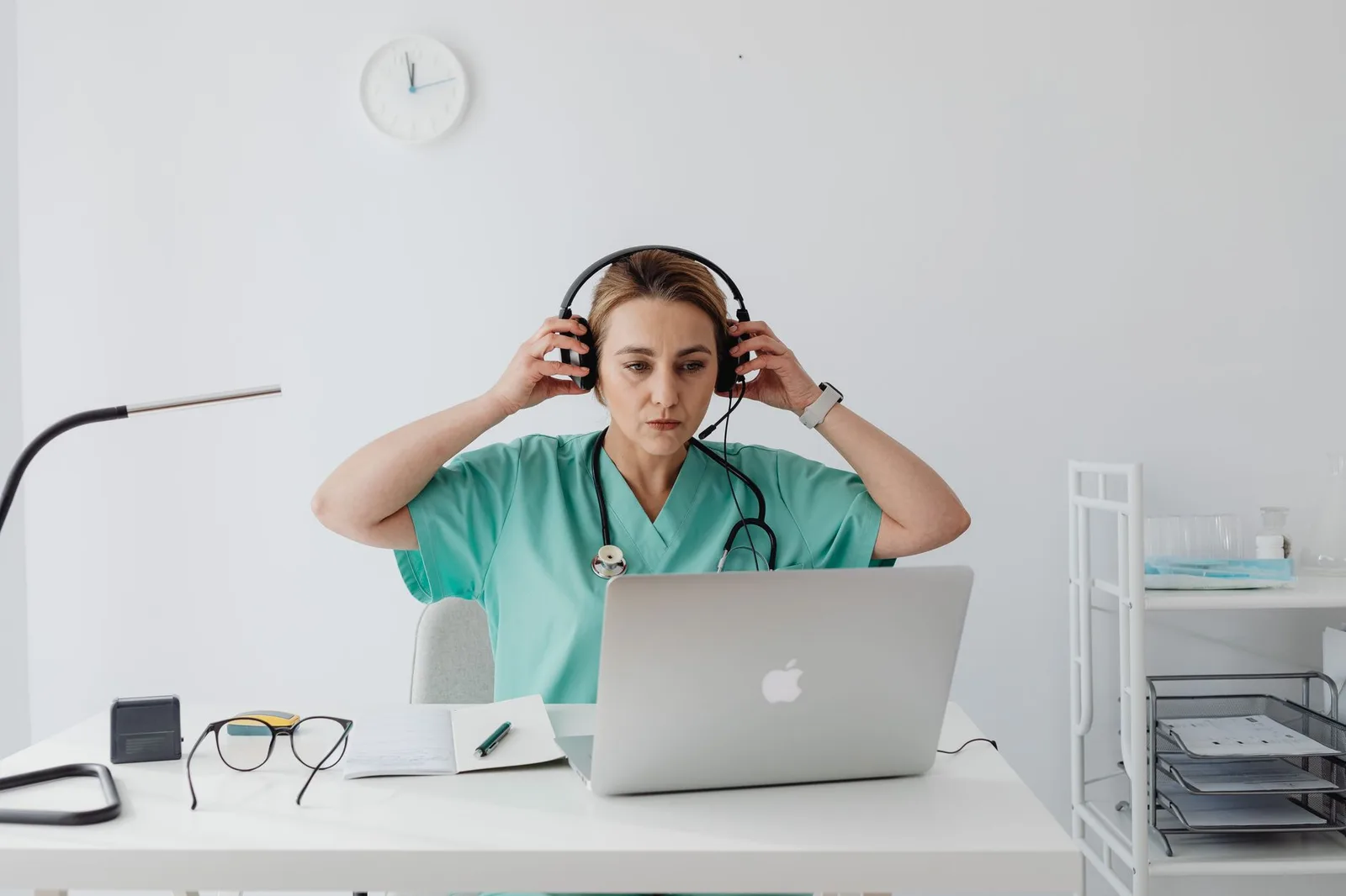 Medical professional in teal scrubs using a laptop and headset, sitting at a desk in an office setting.