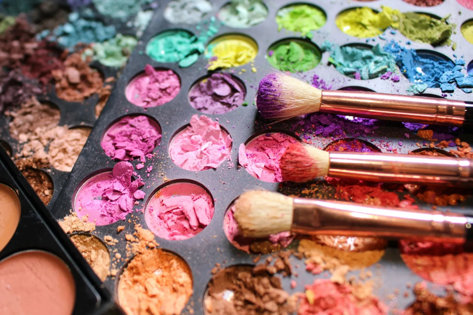 Close-up of colorful crushed makeup palette with three brushes, creating a vibrant artistic mess.