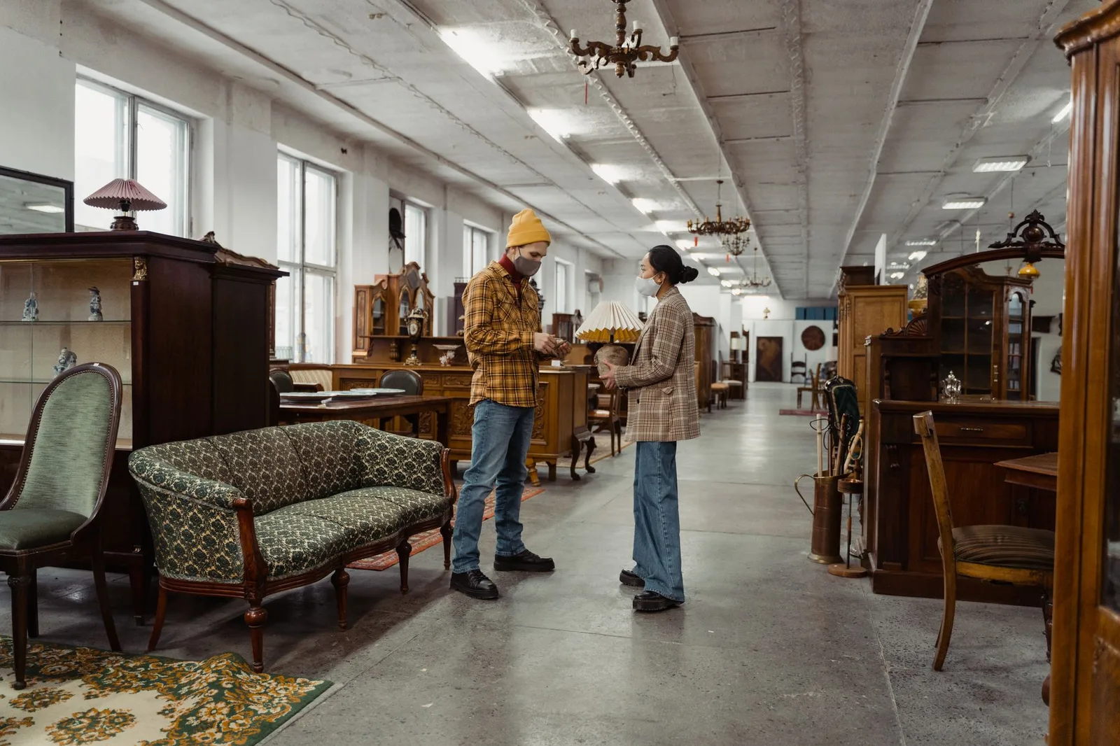 A man and woman wearing masks engage in conversation inside an antique furniture shop.