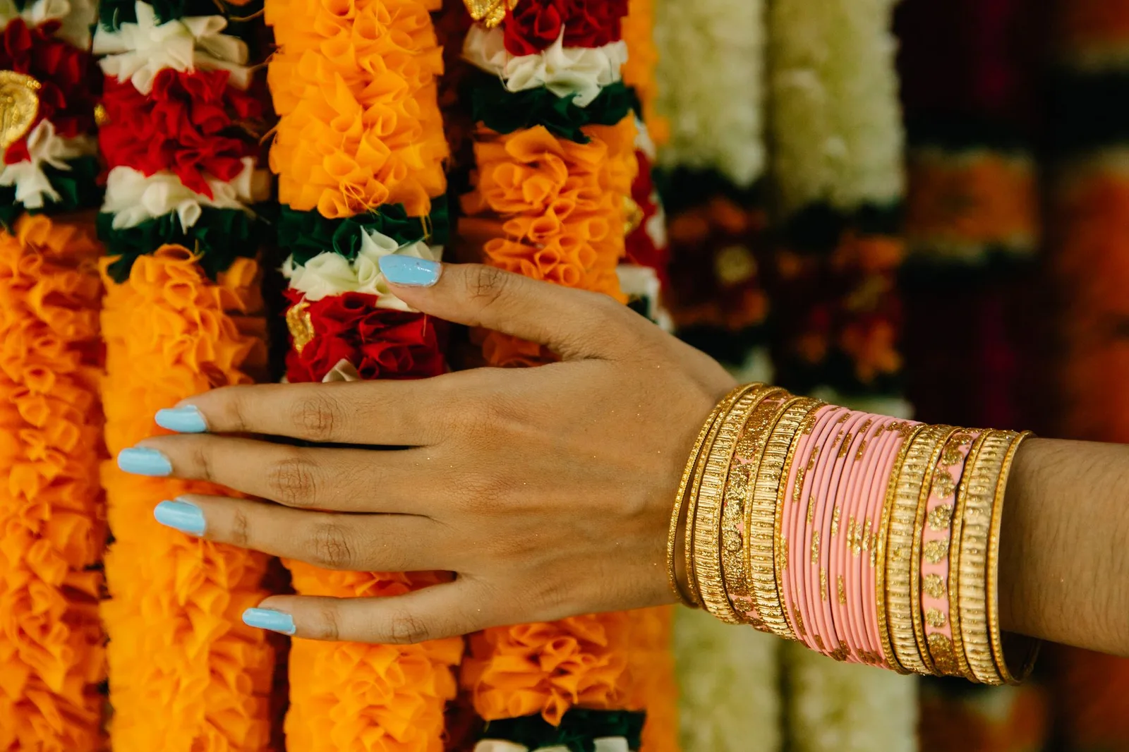 Hand adorned with colorful bangles touching vibrant floral garlands, highlighting cultural richness.